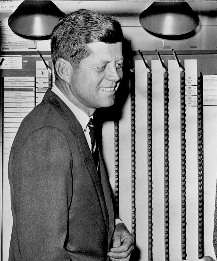 John F. Kennedy smiling beside a voting machine