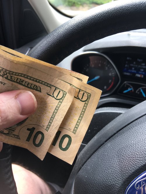 Hand holding a $20 tip in cash at the steering wheel